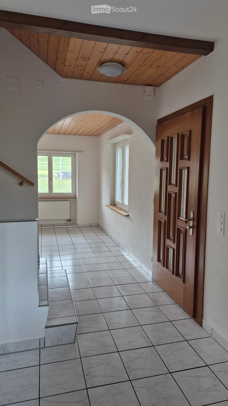 Empty hallway with tiled floor, white walls, wooden door, and arched ceiling with wooden beams. Natural light from windows on the right side.