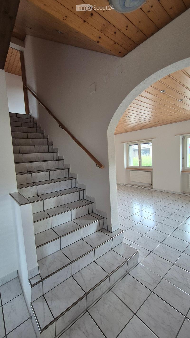 Spacious hallway with a staircase leading upwards, featuring marble tiles and wooden handrails, with a view of a room with large windows and a radiator.