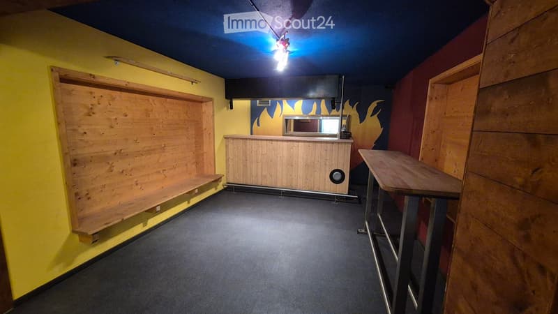 A small, furnished room with a wooden bench, a counter, and a table. The walls are painted in bright yellow and dark blue, with a decorative flame design on one wall.