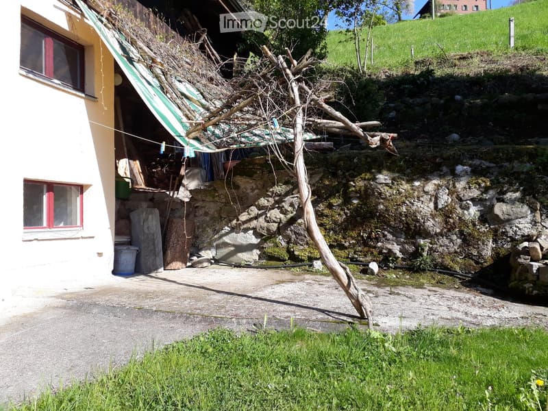 A rustic outdoor area adjacent to a house, featuring a stone wall, a wooden beam, a small covered area with a striped awning, and a grassy slope in the background.