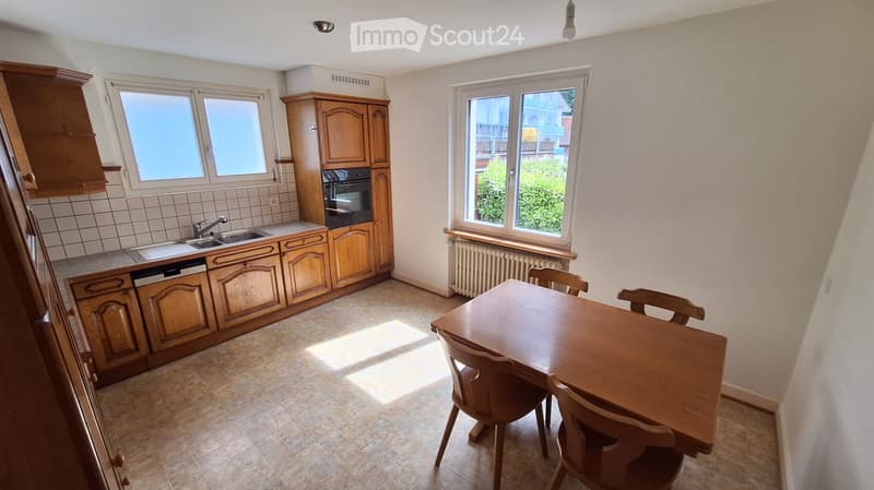 Bright kitchen with wooden cabinets, a sink, an oven, and a dining table with chairs. Natural light streams in from large windows.
