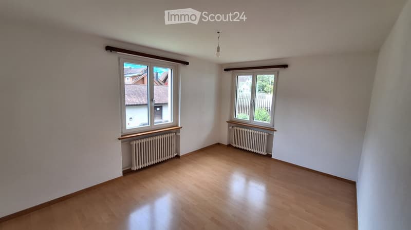 Empty room with wooden flooring, white walls, two windows, radiators, and a ceiling light fixture.