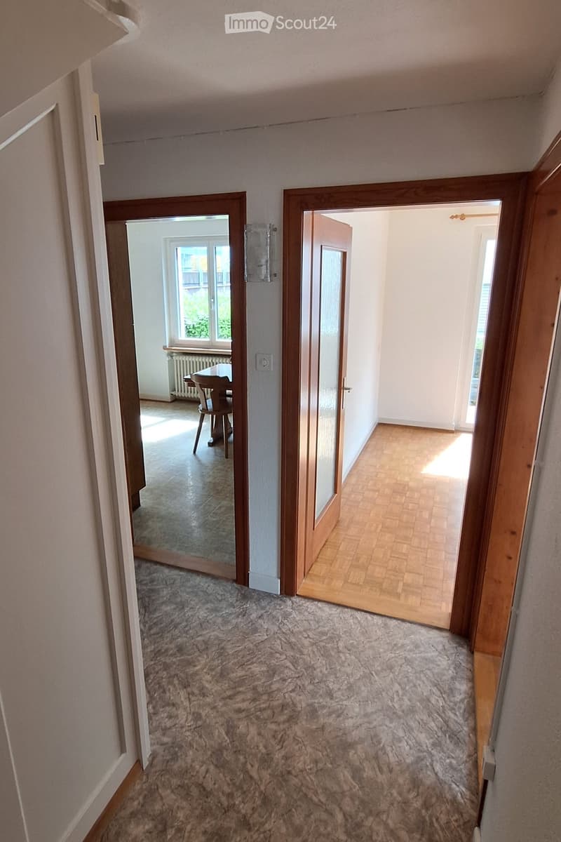 A carpeted hallway with wooden doors leading to adjacent rooms, one of which is a dining area with a table and chairs.
