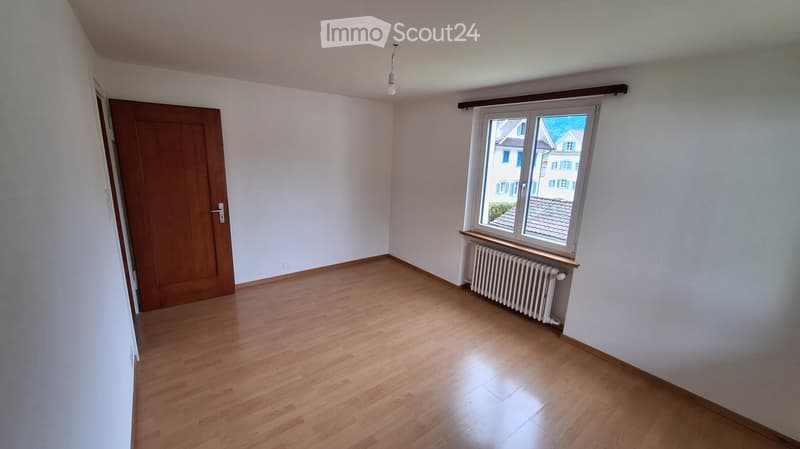 Empty room with wooden flooring, white walls, a window with a radiator below, and a wooden door.