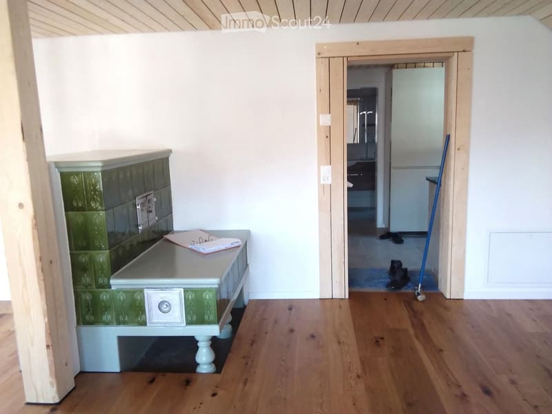A small, well-lit hallway with wooden flooring, a green-tiled bench, and a table with papers on it. A doorway leads to another room with visible furniture.