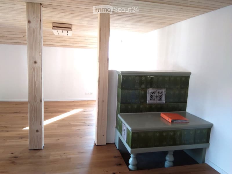 A small, well-lit room with wooden flooring and white walls. A grey table with a green base is positioned against the wall, holding a red book. The room features a wooden pillar and an air conditioning unit on the wall.