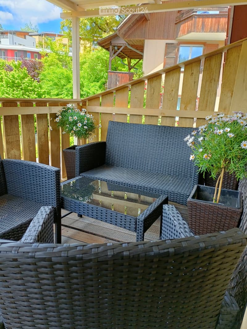 A wooden balcony with wicker furniture, including a sofa, chairs, and a small table, decorated with white flower pots.