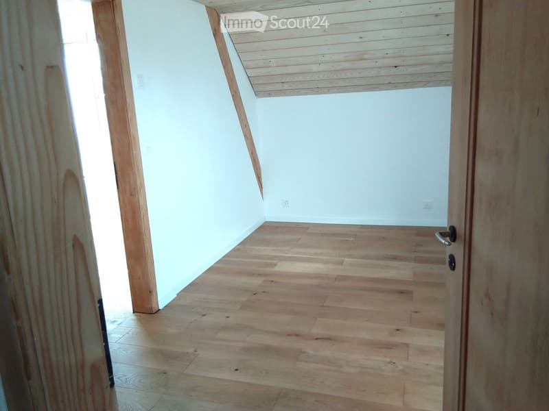 Empty attic space with wooden flooring and slanted wooden ceiling, featuring a doorway leading to another area.