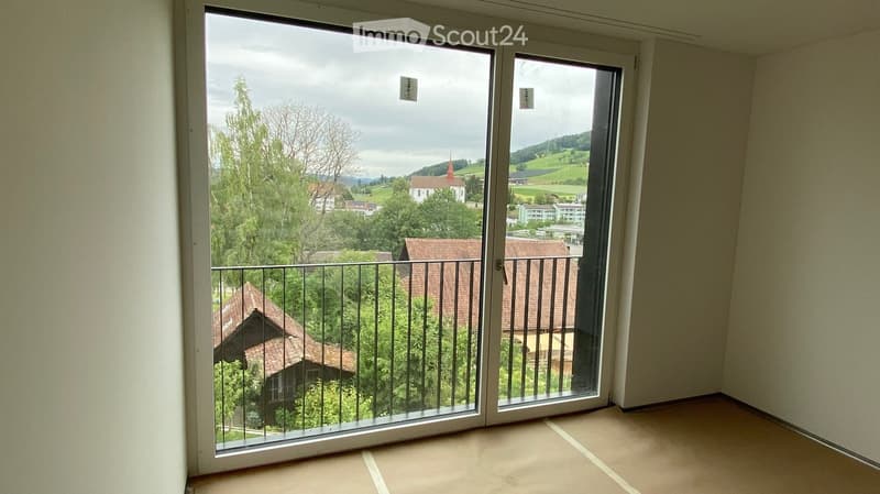 Empty room with large sliding glass doors opening to a balcony, overlooking a scenic countryside with houses and trees.