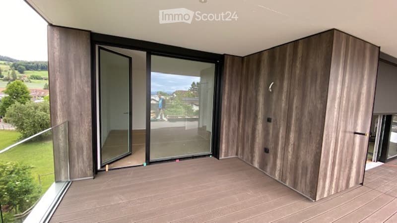 Modern balcony with large glass doors, wooden flooring, and wooden panel walls, offering scenic views of a green landscape.