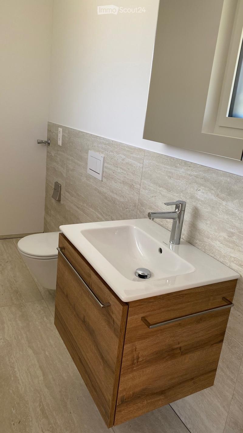 Modern bathroom with a white sink, chrome faucet, wooden cabinet, and a toilet. Light-colored tiles and a large mirror are visible.
