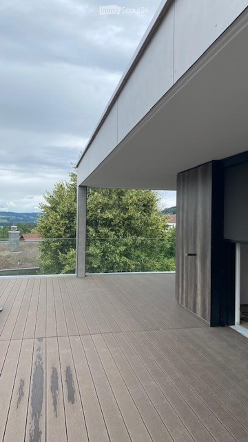 Modern wooden deck terrace with glass railing, overlooking greenery and distant hills.