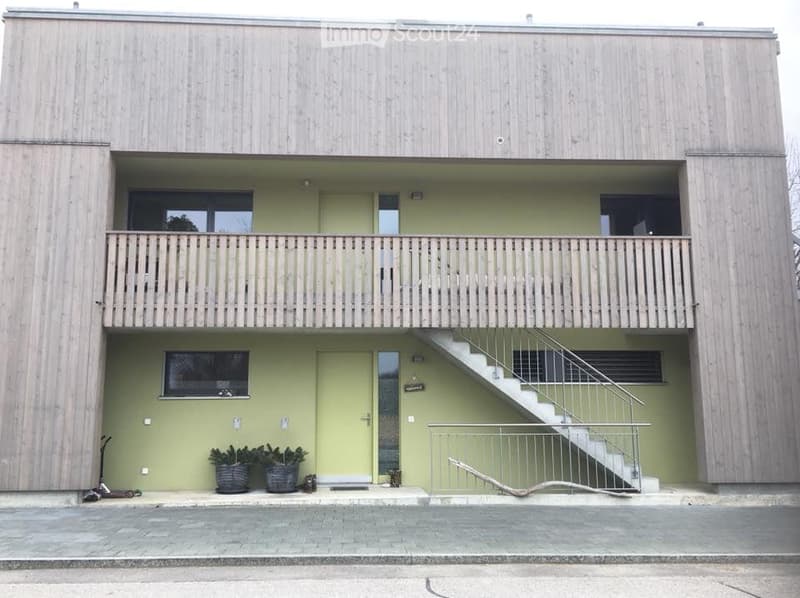 Modern two-story building with light wood siding, green lower facade, balcony with wooden railing, staircase leading to upper level, and potted plants near entrance.