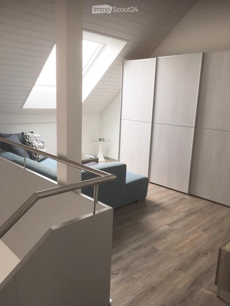 Modern, loft-style living area with wooden flooring, white walls, and skylight windows. Features a blue sofa, matching ottoman, small round table, and decorative pillows.