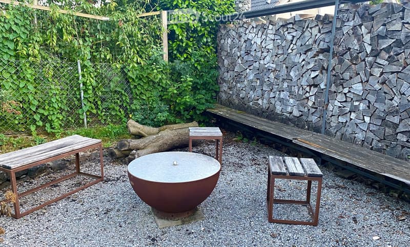 Garden area with gravel ground, wooden benches, a metal fire pit, a log pile, and a wooden fence with greenery.