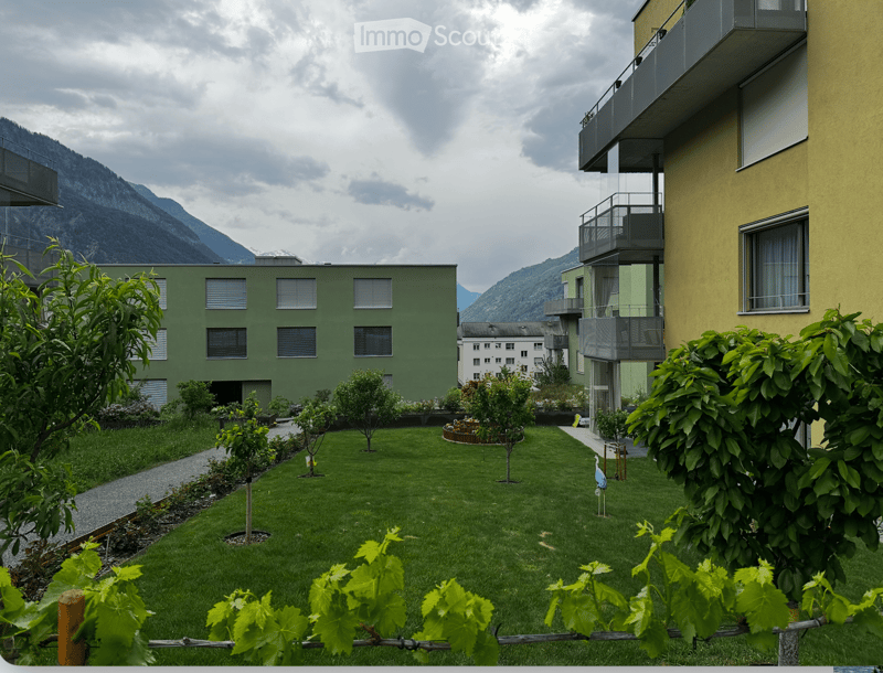 Well-maintained garden area with green grass, small trees, and shrubs, surrounded by modern apartment buildings and a scenic mountain backdrop.