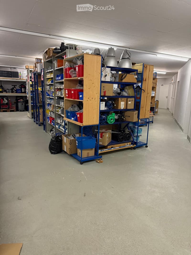Empty warehouse or storage area with concrete floor, metal shelving units, and various storage items including boxes, tools, and equipment.
