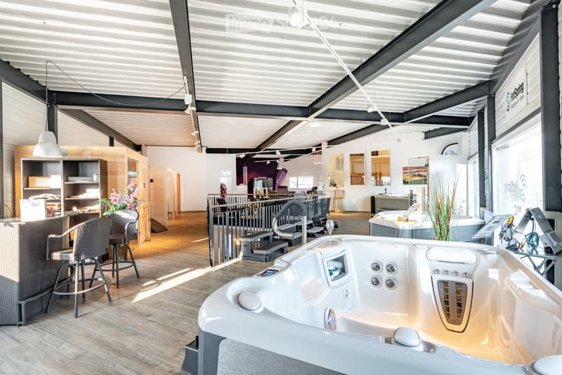Modern showroom with a large hot tub in the foreground, surrounded by seating areas, a counter, and display cabinets. The space features exposed metal beams and a sleek, contemporary design.
