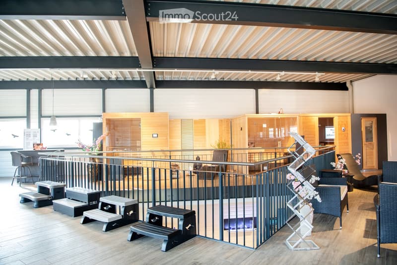 Modern, open-plan space with wooden flooring, metal railing, and various seating options including stools and chairs. Features a display stand with brochures and a small table with a flower vase.