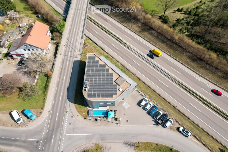 Aerial view of a modern building with a solar panel roof, surrounded by a parking lot with several cars, a small pool, and a nearby road with traffic.