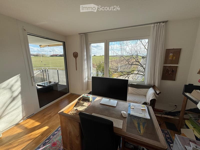 Bright room with wooden flooring, large windows, a sofa, a desk with a computer setup, and artwork on the walls.