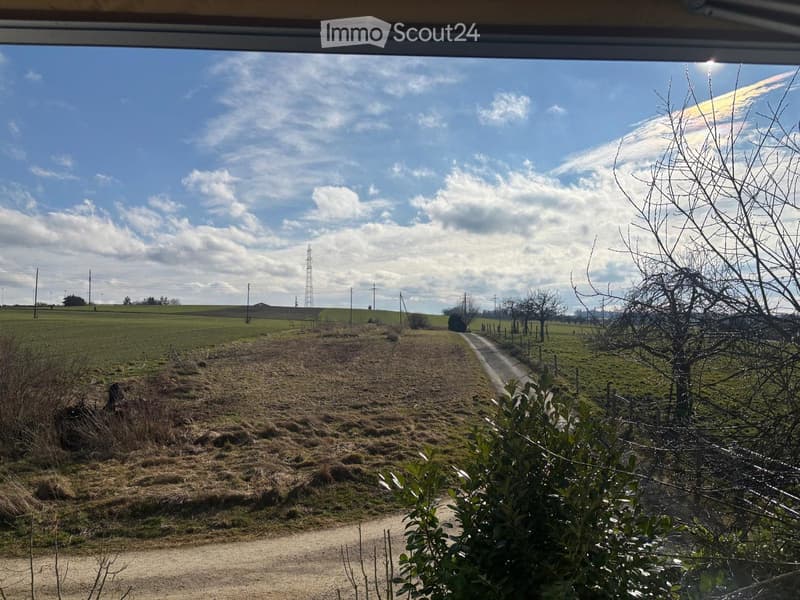 View from a window showing a rural landscape with fields, a dirt road, and scattered trees under a partly cloudy sky.
