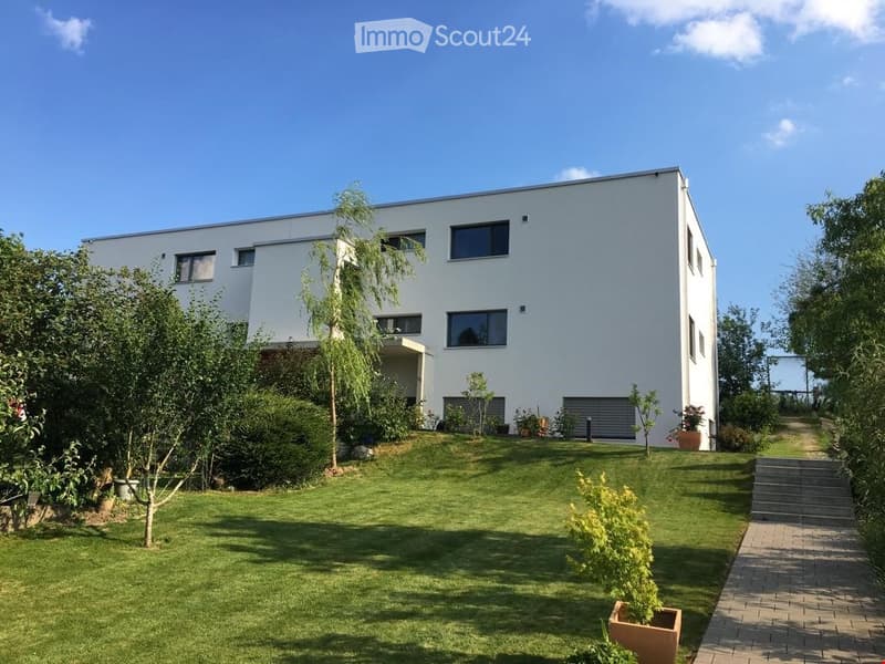 Two-story house with modern architectural design, featuring a white exterior, large windows, and a balcony on the second floor. The property is situated on a sloping terrain with a driveway leading to the entrance. The garden includes lush greenery, shrub