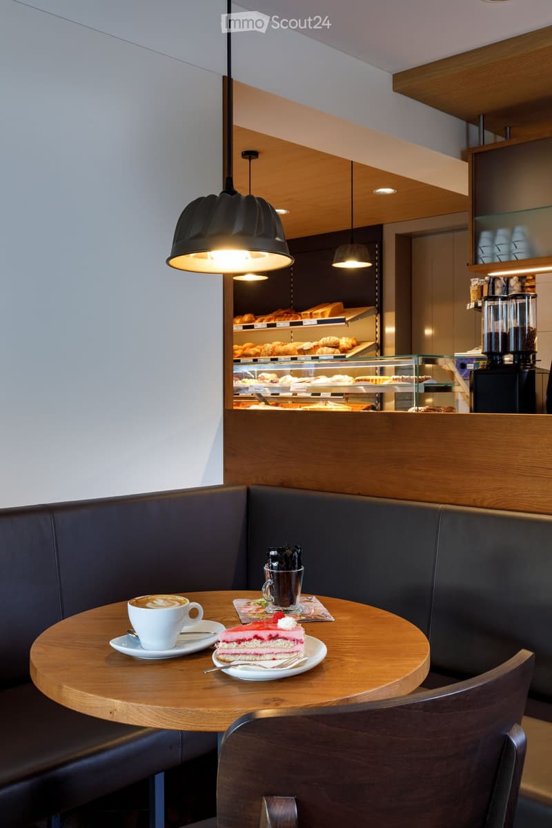 A small round table with a white cup and saucer, a plate with a slice of cake, and a glass of coffee. A black leather booth is on the left side, and a wooden chair is on the right side. A lamp is hanging from the ceiling, and a glass display case with bre