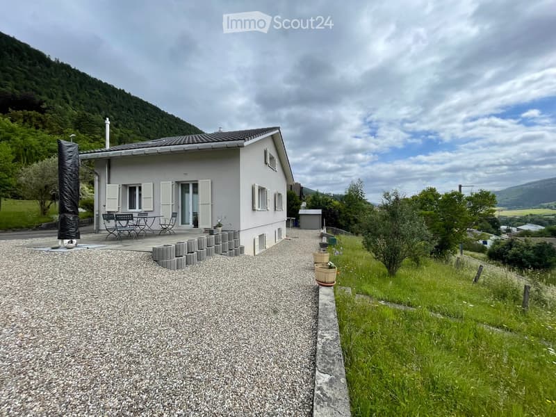 Single-story house with white shutters, gravel driveway, outdoor seating area, and scenic mountain backdrop.