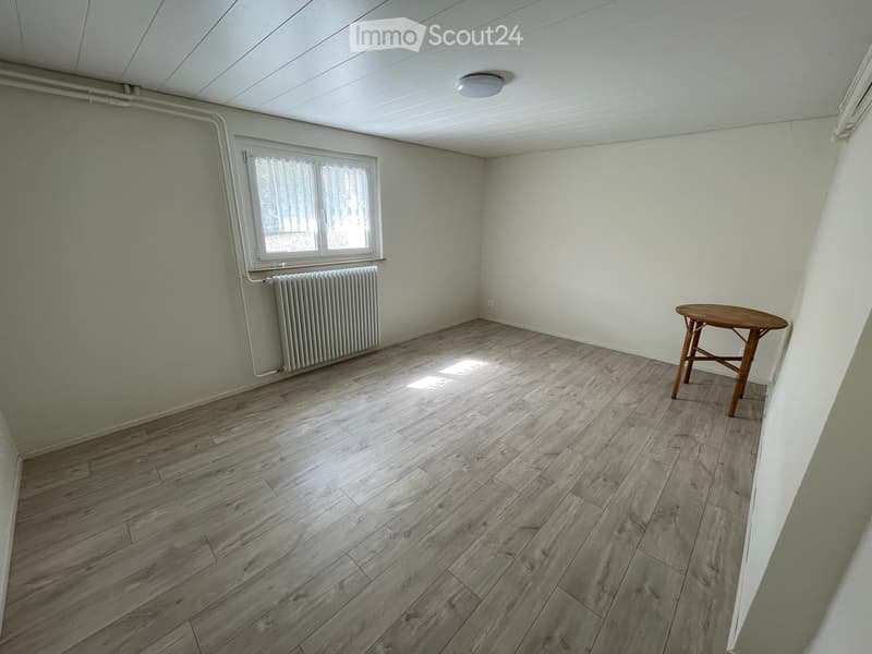 Empty room with wooden flooring, a single window, radiator, and a small wooden stool.