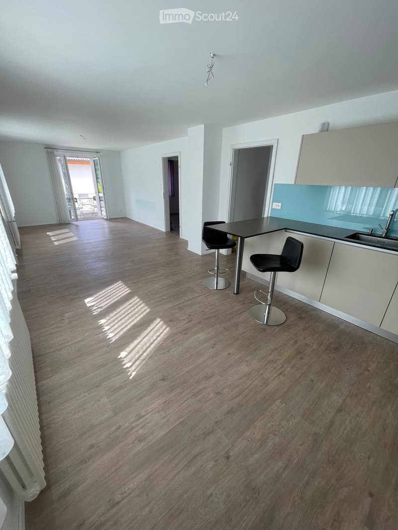 Open-plan living and dining area with wooden flooring, white walls, and large windows. Includes a small dining table with two stools and a modern kitchen area with a sink and cabinets.