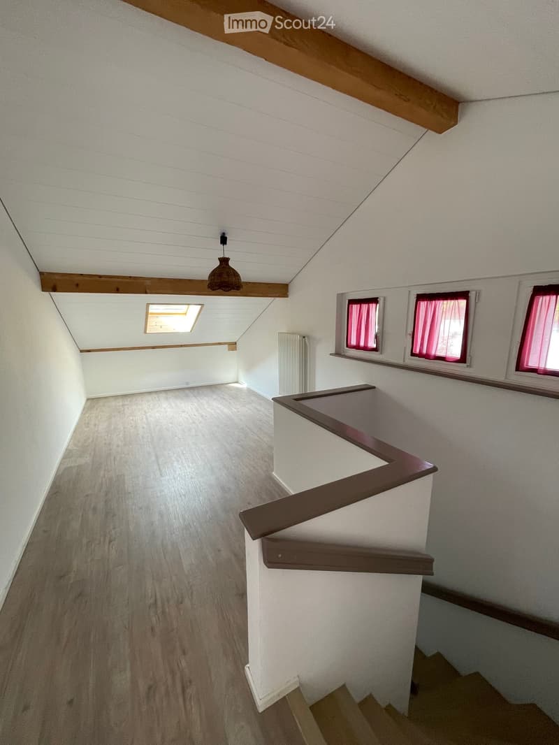 Empty hallway with wooden flooring, white walls, and a staircase leading upwards. Features include a hanging light fixture, a small skylight, and windows with pink curtains.