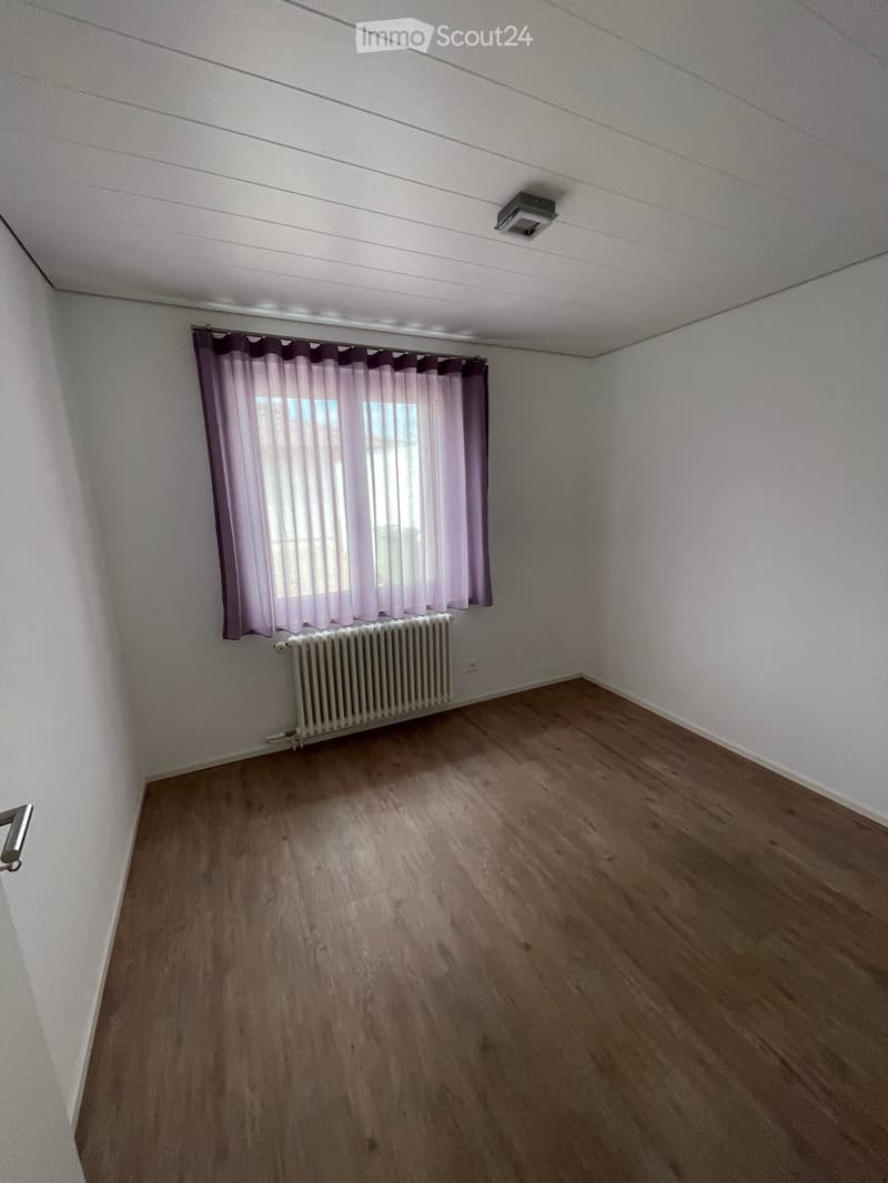Empty room with wooden flooring, white walls, a window with purple curtains, and a radiator below the window.