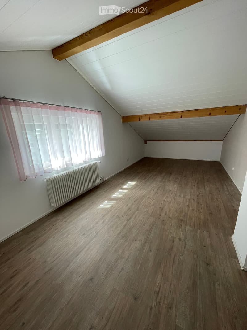Empty attic room with wooden flooring, white walls, and a slanted ceiling. Features a window with pink curtains and a radiator below it.
