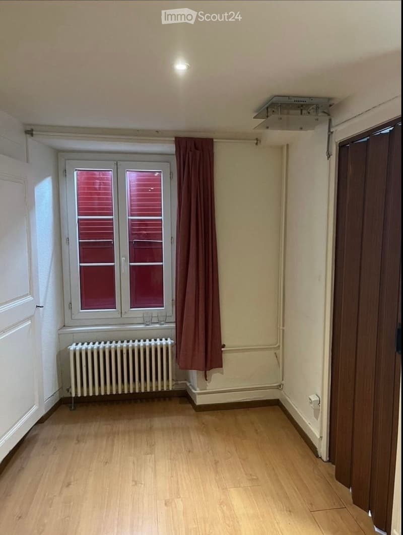 Empty room with wooden flooring, white walls, a window with red blinds, a radiator, and a door.
