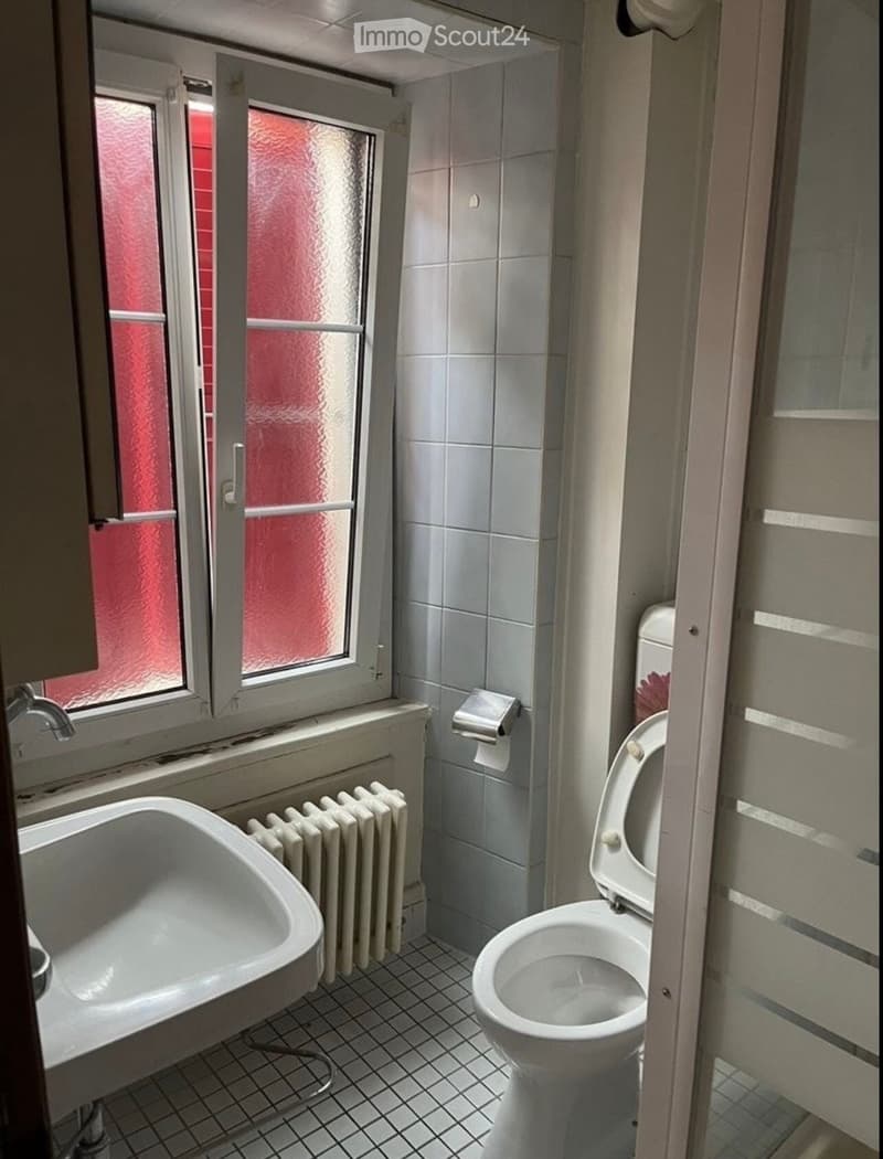 Bathroom with a toilet, sink, radiator, and toilet paper holder. The room has tiled walls and floor, and a window with frosted glass.