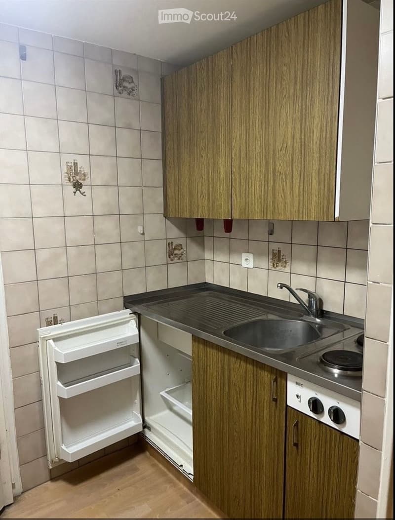 Compact kitchen with wooden cabinets, a sink, a stove, and an open refrigerator. The walls are tiled with decorative tiles.