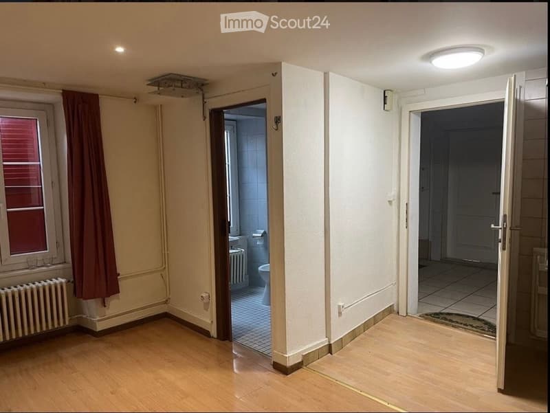 Empty room with wooden flooring, white walls, and a window with red blinds. Adjacent rooms visible through open doors include a bathroom and another room with tiled flooring.