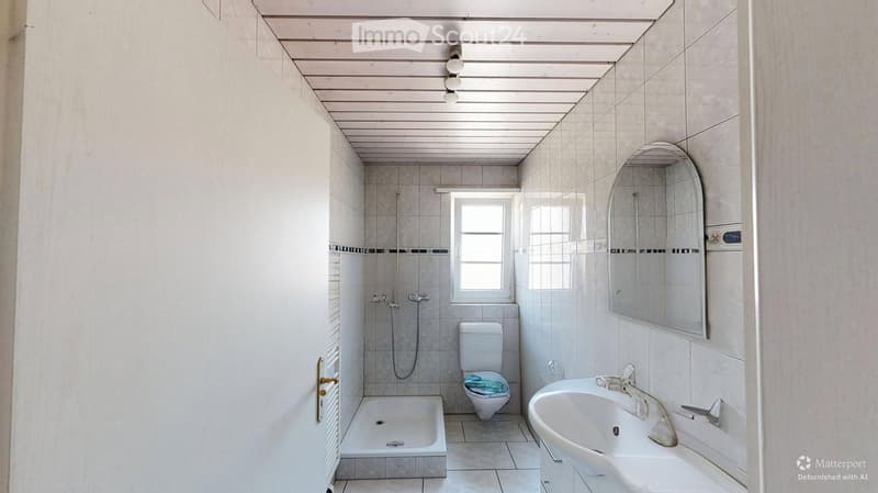 Bathroom with white tiles, a toilet, a sink, a shower, and a mirror. The room has a window and is well-lit.