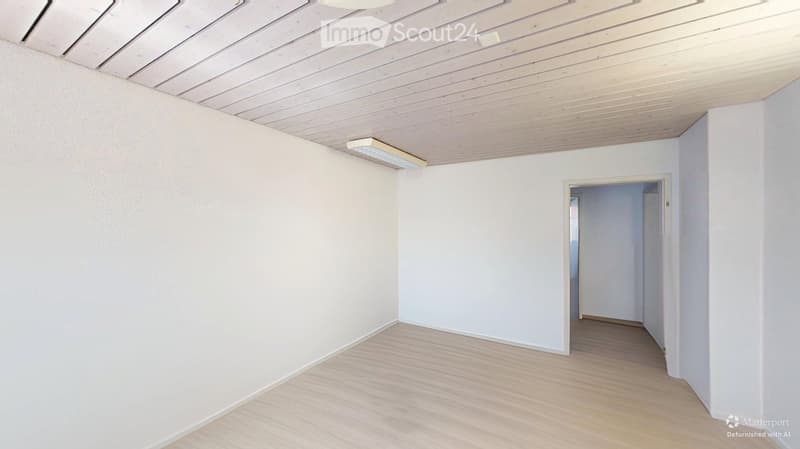 Empty room with light wooden flooring, white walls, and a wooden ceiling. A doorway leads to another room.