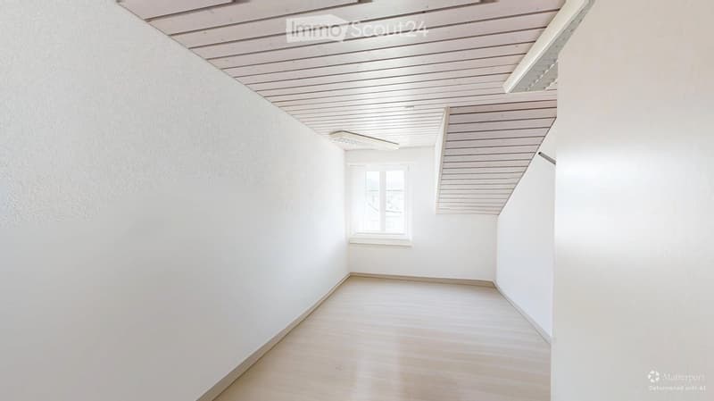 Empty room with white walls, wooden floor, and a single window. The ceiling has a slanted, paneled design.