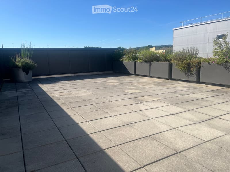 A modern rooftop terrace with a tiled concrete floor, grey planters with greenery, and a low metal railing. The area is clean and spacious, with a clear blue sky in the background.