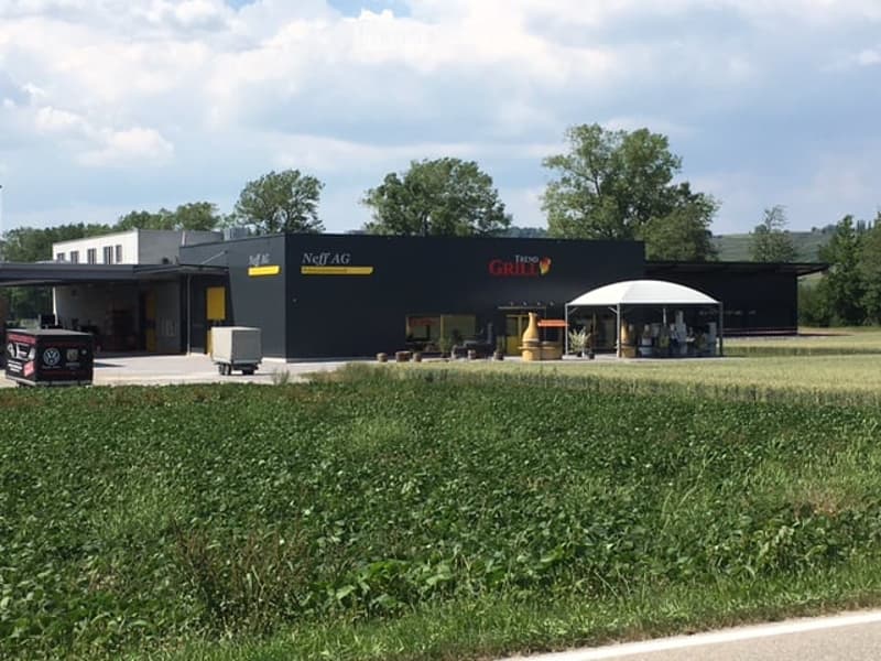 Modern commercial building with a black facade, signage for "Neff AG" and "Tenne Grill", outdoor seating area, and a grassy lawn in front.