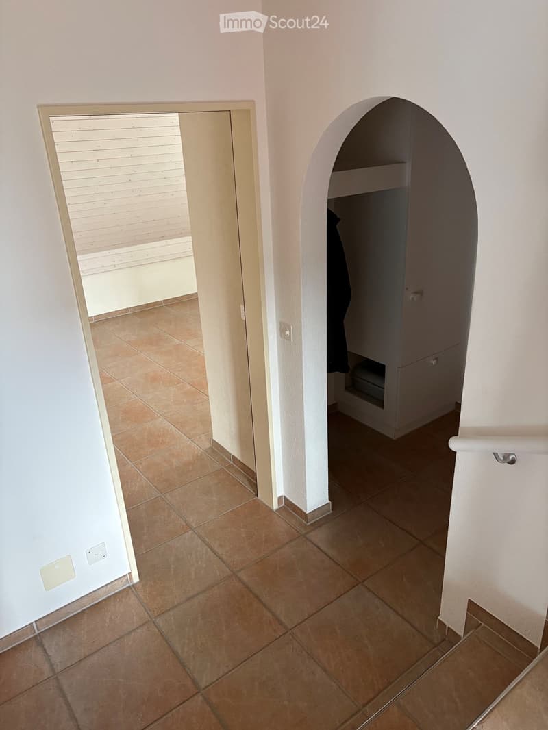 A hallway with beige tiled flooring, white walls, and an arched doorway leading to a closet with a single hanging garment visible. A sliding glass door with blinds is visible on the left side.