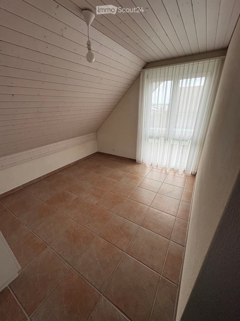 Empty room with wooden slat ceiling, tiled floor, and large window with sheer curtains.