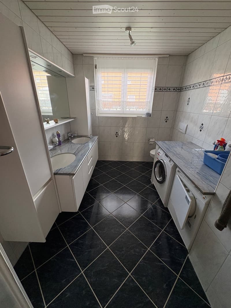 Bathroom with dark tiled floor, white cabinets, double sink, washing machine, dishwasher, and a window with blinds.