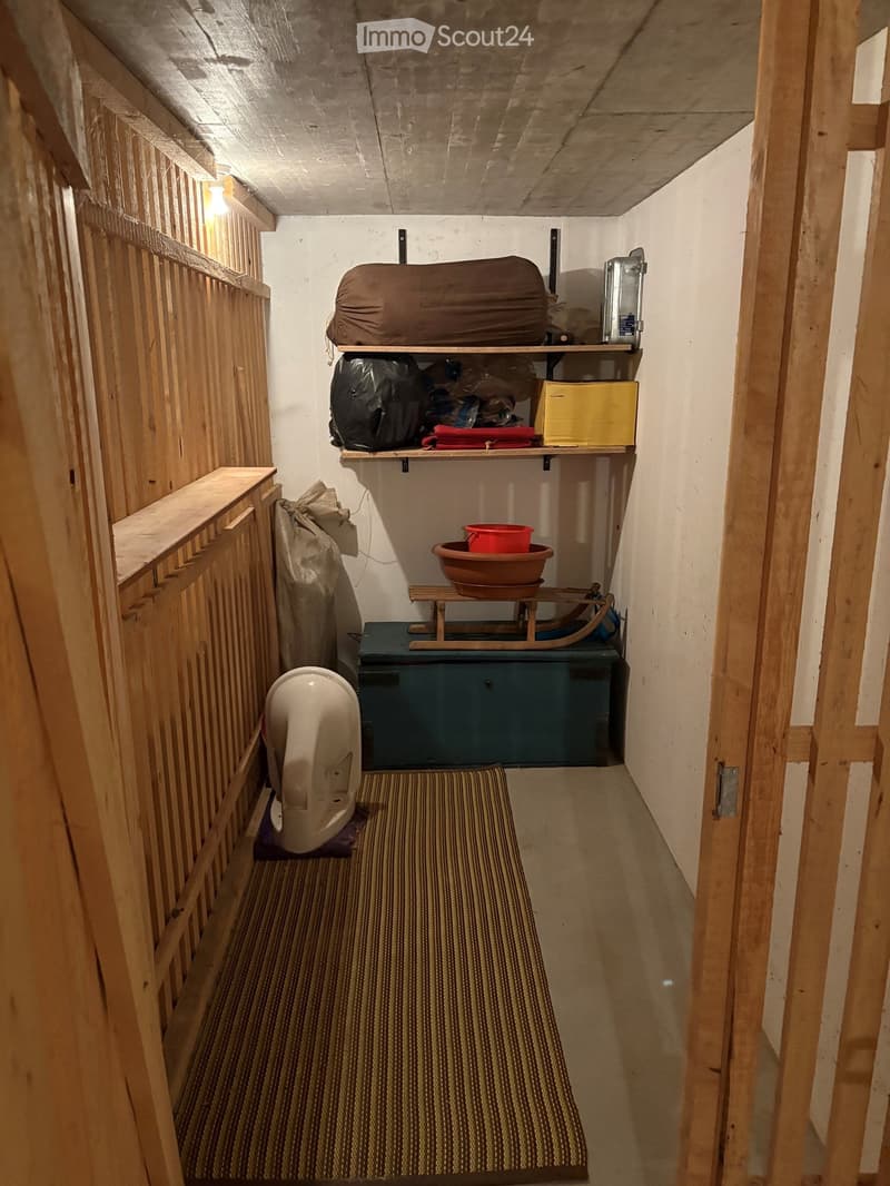 A narrow, wooden-paneled room with shelves holding various items, including a mattress, boxes, and a stool with a pot on top. A small rug covers part of the floor, and a white object is placed near the left wall.