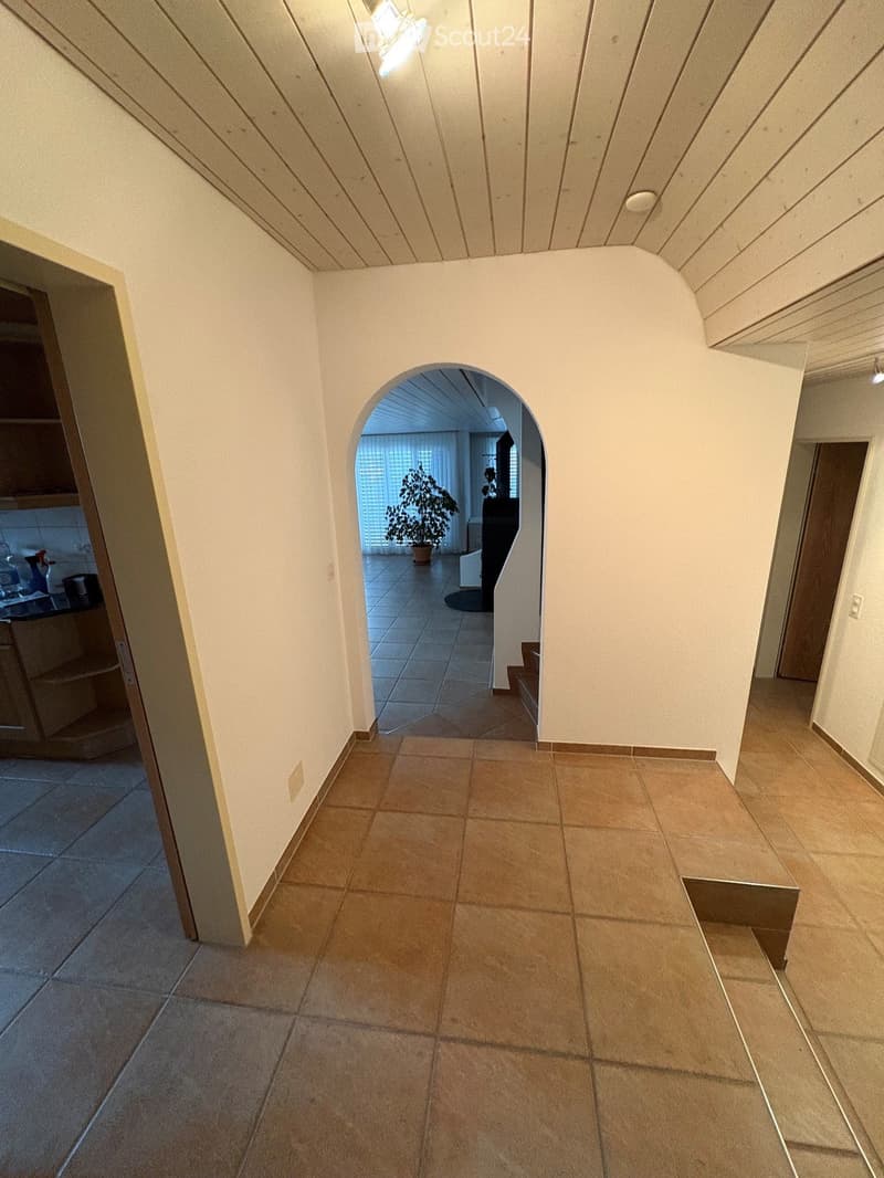 A well-lit hallway with tiled flooring, white walls, and wooden ceiling beams. An arched opening leads to another room with a visible potted plant and a staircase.