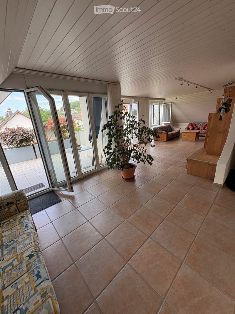 Bright living room with large windows and sliding doors leading to a balcony, featuring a patterned sofa, a wooden coffee table, and a potted plant.
