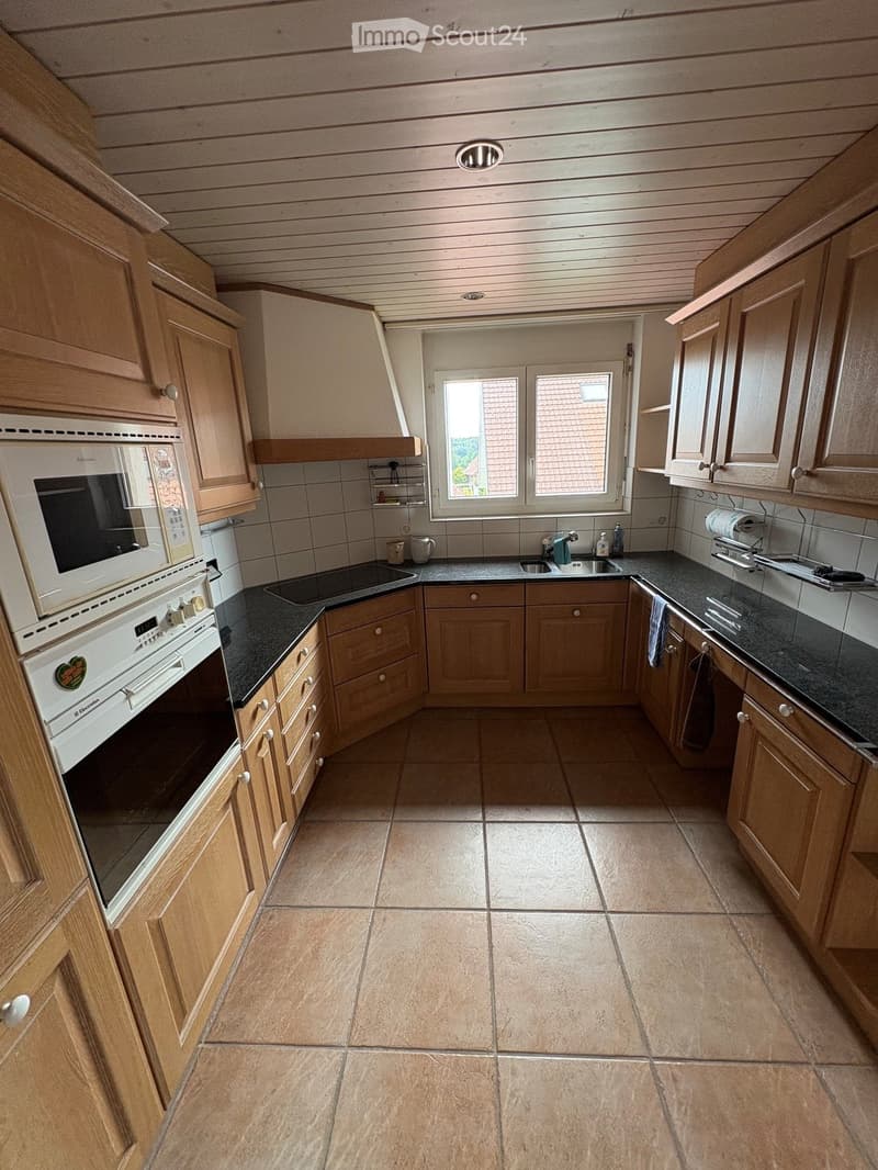 A well-lit kitchen with wooden cabinets, a black countertop, and a tiled floor. The kitchen features an oven, microwave, sink, and various small appliances.